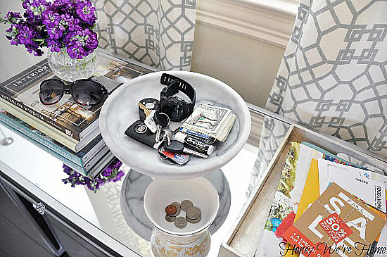 Tray filled with change, money, watch, keys, chapstick. On table in front of window.