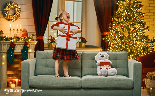 Girl holding a Christmas gift, standing on a sofa and screaming. Christmas tree and poinsettas in background