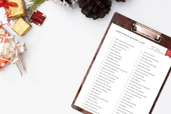 christmas bucket list printable on clipboard on white desk surrounded by christmas gifts and pinecones