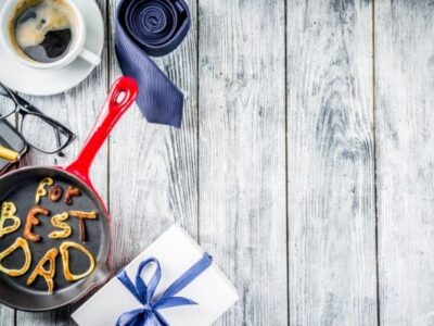 Pancakes that are made to say "best dad" in frying pan, blue tie, coffee mug, present on white wooden table