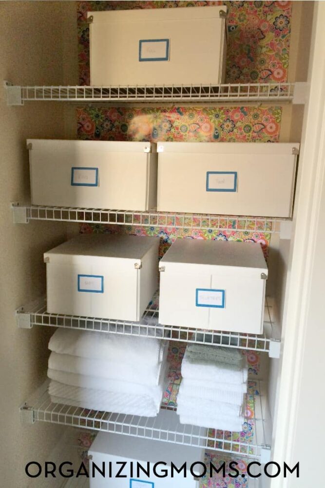 White boxes used for linen closet organization in a linen closet with white wire shelves and floral wallpaper