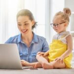An organized mom sitting at a table using a laptop with preschool girl next to her