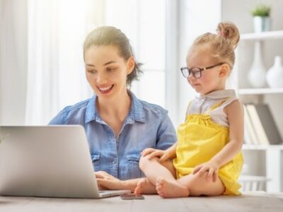 An organized mom sitting at a table using a laptop with preschool girl next to her
