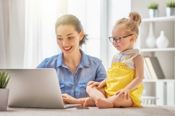 An organized mom sitting at a table using a laptop with preschool girl next to her