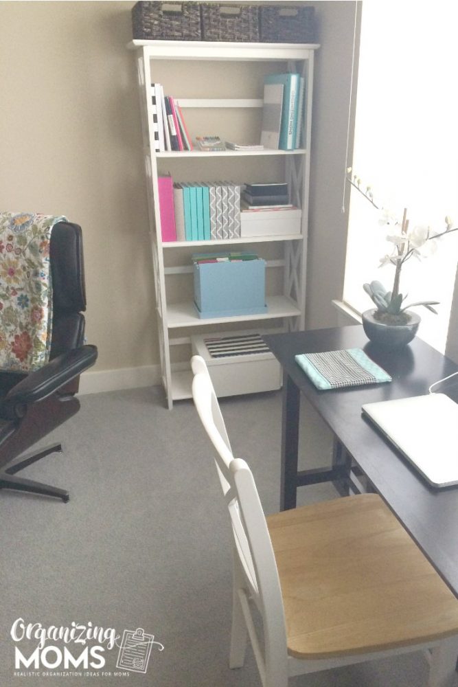 A desk with a computer and a chair in a room. Organized office bookshelf.