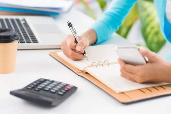 Woman doing quarterly planning with planner, cell phone and calculator at desk with computer.