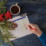 man scheduling holiday activities on calendar with christmas tree branch, ornaments, and coffee on the table