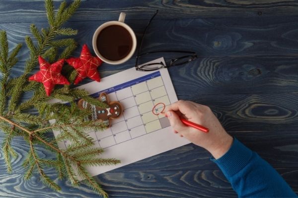 Man scheduling holiday activities on calendar with christmas tree branches, decorations and coffee on table