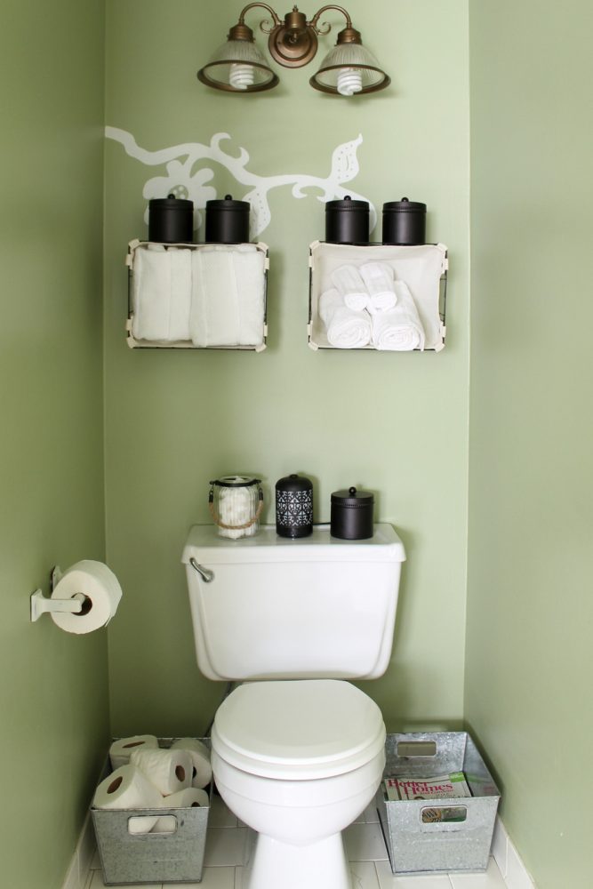 baskets anchored to wall above toilet holding towels. Various black containers used to hold toiletries.