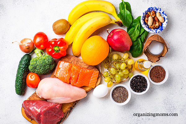 Healthy food items on a white counter. Salmon, chicken, orange, apple, grapes, broccoli and more. 