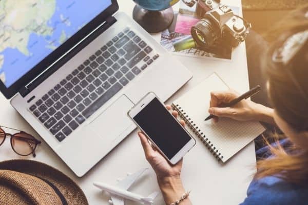 woman writing in notebook brain dump looking at phone in front of computer